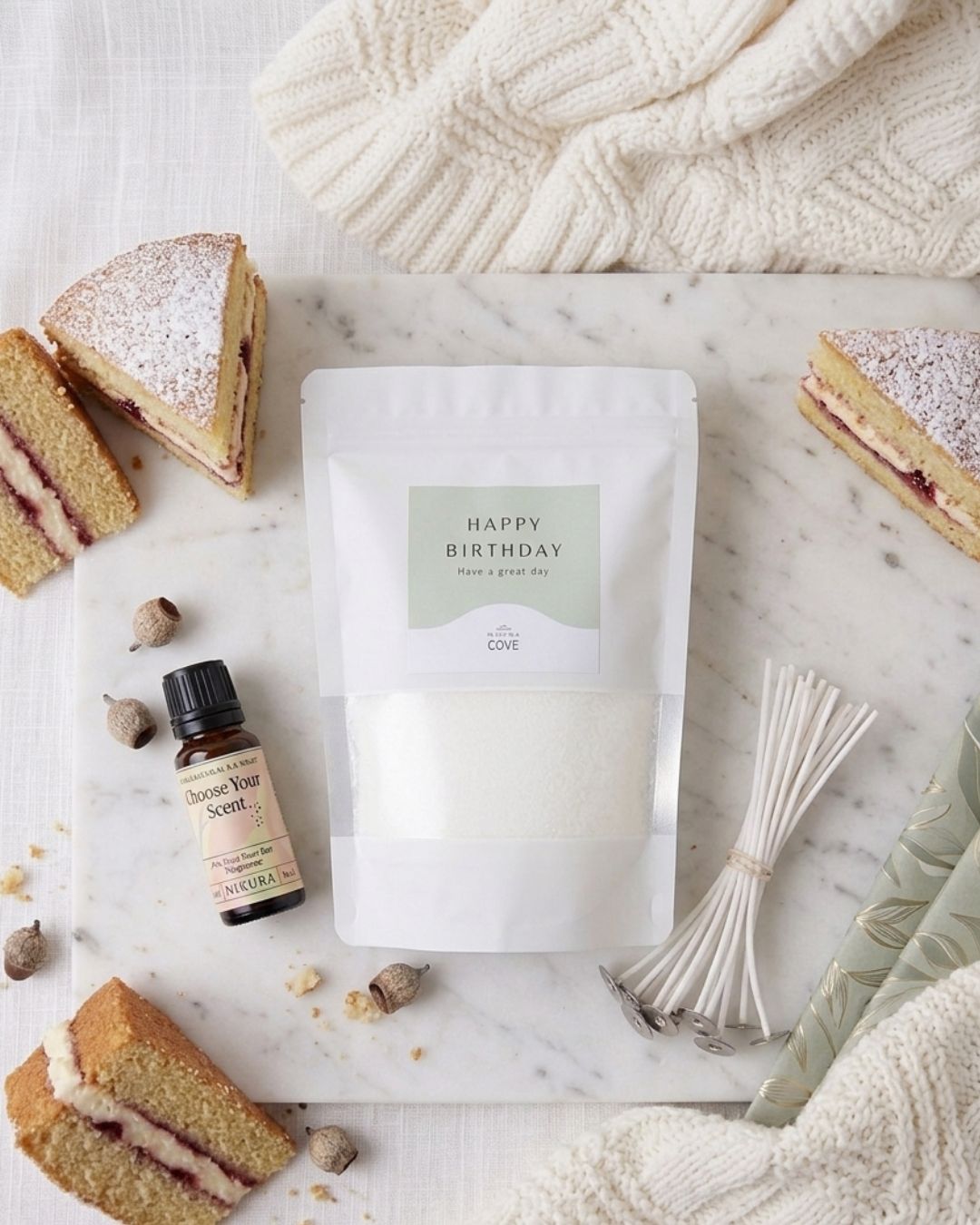 A flat-lay of a white birthday candle gift set on a marble board, surrounded by cake slices, a small bottle of scent and cotton wicks.