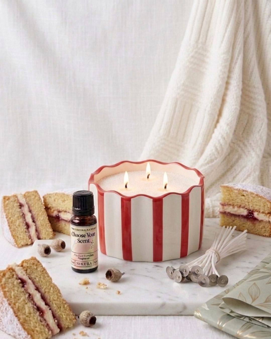 A three-wick scented candle in a white ceramic pot with red vertical stripes sits on a marble surface, surrounded by slices of layered cake,  a bottle of essential oil and a bundle of wicks.