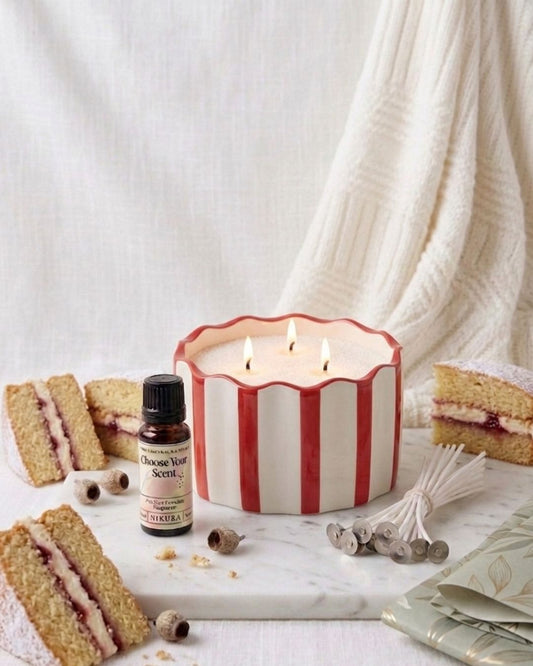 A three-wick scented candle in a white ceramic pot with red vertical stripes sits on a marble surface, surrounded by slices of layered cake,  a bottle of essential oil and a bundle of wicks.