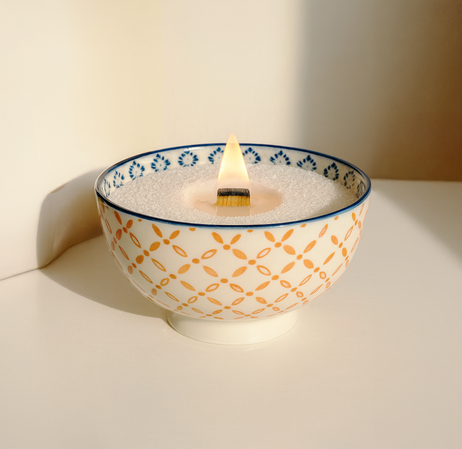 Sunlight casting a warm shadow on a DIY refillable candle. The setup features a wide-flame wooden wick and plant-based wax sand inside a decorative blue and orange bowl by White Cove UK