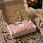 A beautifully packaged Mother's Day eco-friendly candle gift set by White Cove. The open mailer box shows pink tissue paper sealed with a logo sticker, pre-tabbed wicks, and two rustic coasters, set against a cozy fireplace