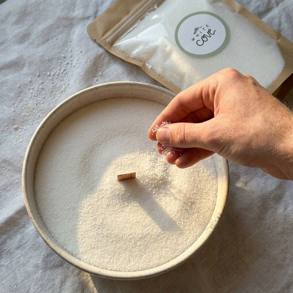 A hand sprinkles white candle sand, poured into a shallow dish filled with white candle sand, in the background on a light fabric surface.