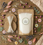 A flat lay of the White Cove Mother's Day Candle Sand Gift Bundle on a rustic wooden table, featuring a kraft pouch of vegetable-based wax sand, a bundle of cotton wicks, and woven coasters, surrounded by dried pink and purple florals