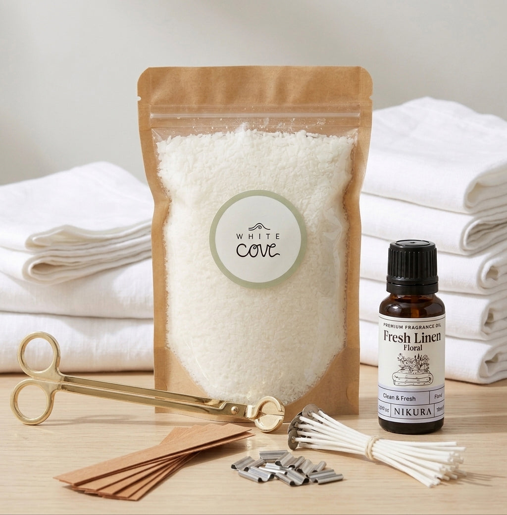 A complete White Cove UK refillable candle sand bundle on a wooden surface. The kit includes a kraft pouch of vegetable-based wax granules, a gold wick trimmer, cotton wicks, wooden wicks with metal bases, and Nikura Fresh Linen essential oil, styled in front of folded white towels