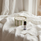 A green and white striped ceramic bowl repurposed into a three-wick candle using White Cove UK refillable candle sand. A 10ml bottle of Nikura Fresh Linen essential oil sits beside it on draped white linen fabric near a bright window
