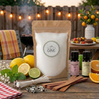 Outdoor summer dining setup featuring the White Cove UK sustainable candle making kit. This lifestyle hero shot showcases the refillable candle sand bundle paired with Lemon and Geranium fragrance oils, arranged on a patio table for an al fresco BBQ setting with fresh fruit, food skewers, and warm festoon lights