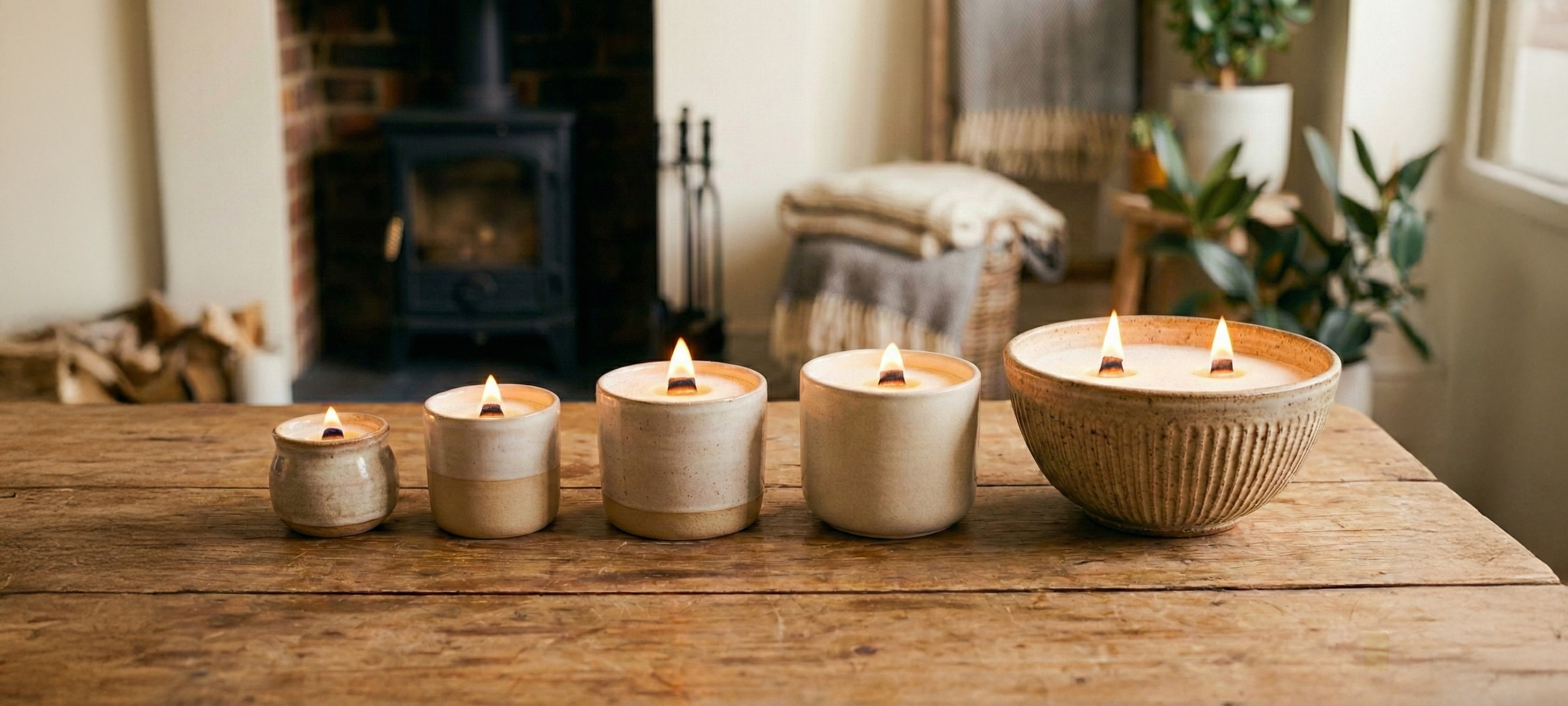 A row of five neutral ceramic vessels repurposed into candles using White Cove UK refillable candle sand. The paraffin-free, vegetable-oil-based wax granules are lit with wooden wicks, displayed on a rustic wooden table in a cozy living room featuring a wood-burning stove