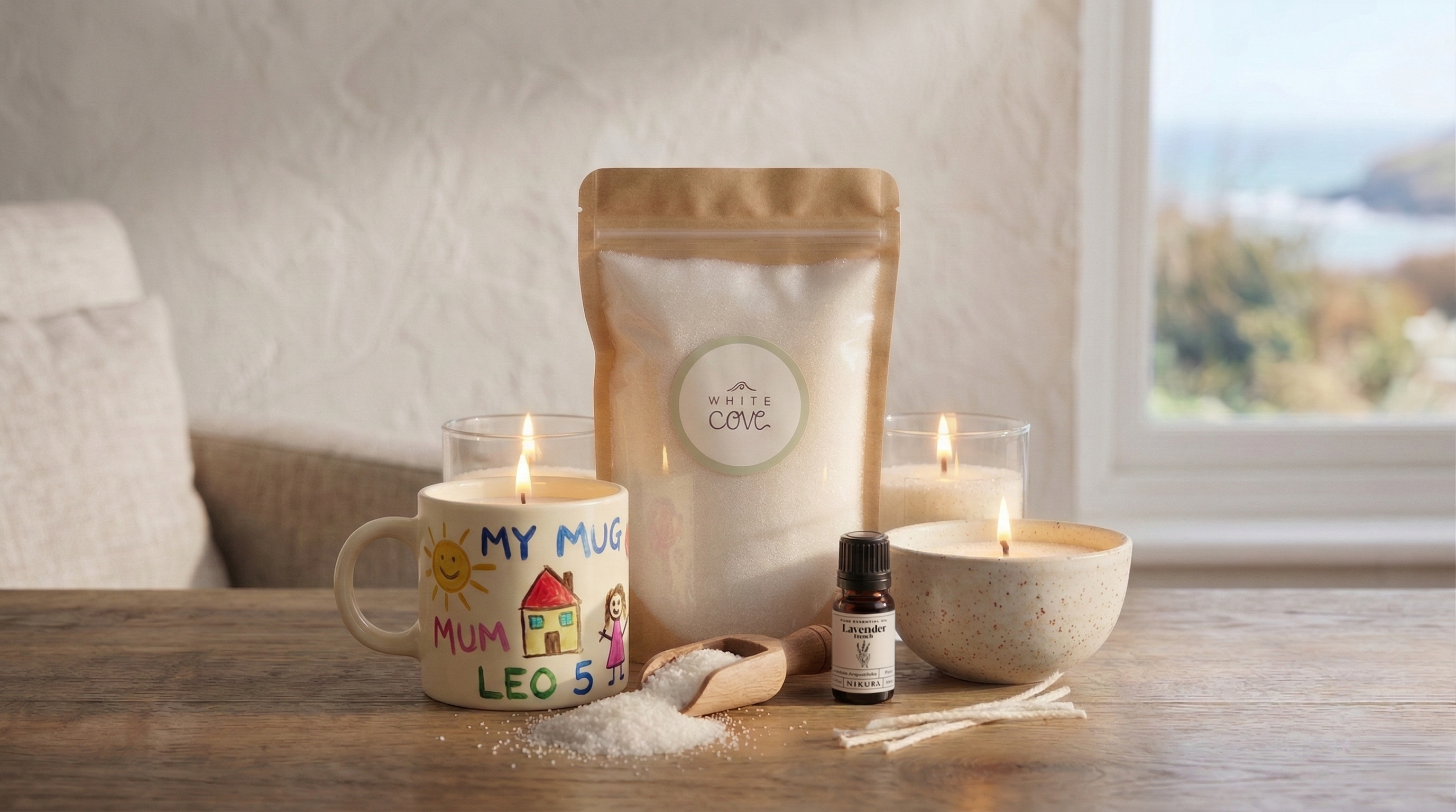 A White Cove refillable candle sand pearled wax display featuring a standing wax pouch, Nikura essential oil, and lit candles in a child's hand-painted mug and a ceramic bowl. Styled on a wooden table with a coastal Dorset sea view in the background