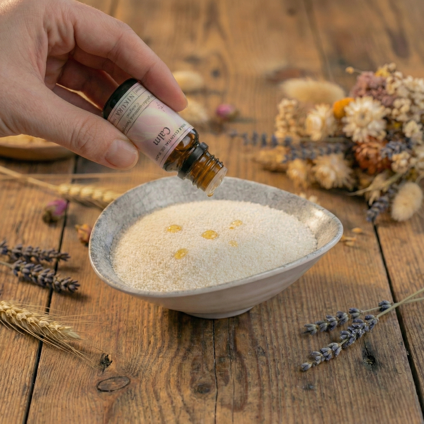 Customising White Cove UK's plant-based candle sand with premium Nikura essential oils. A person adds scent to the white wax granules in a ceramic bowl, styled with dried botanicals for a calm, minimal home fragrance ritual